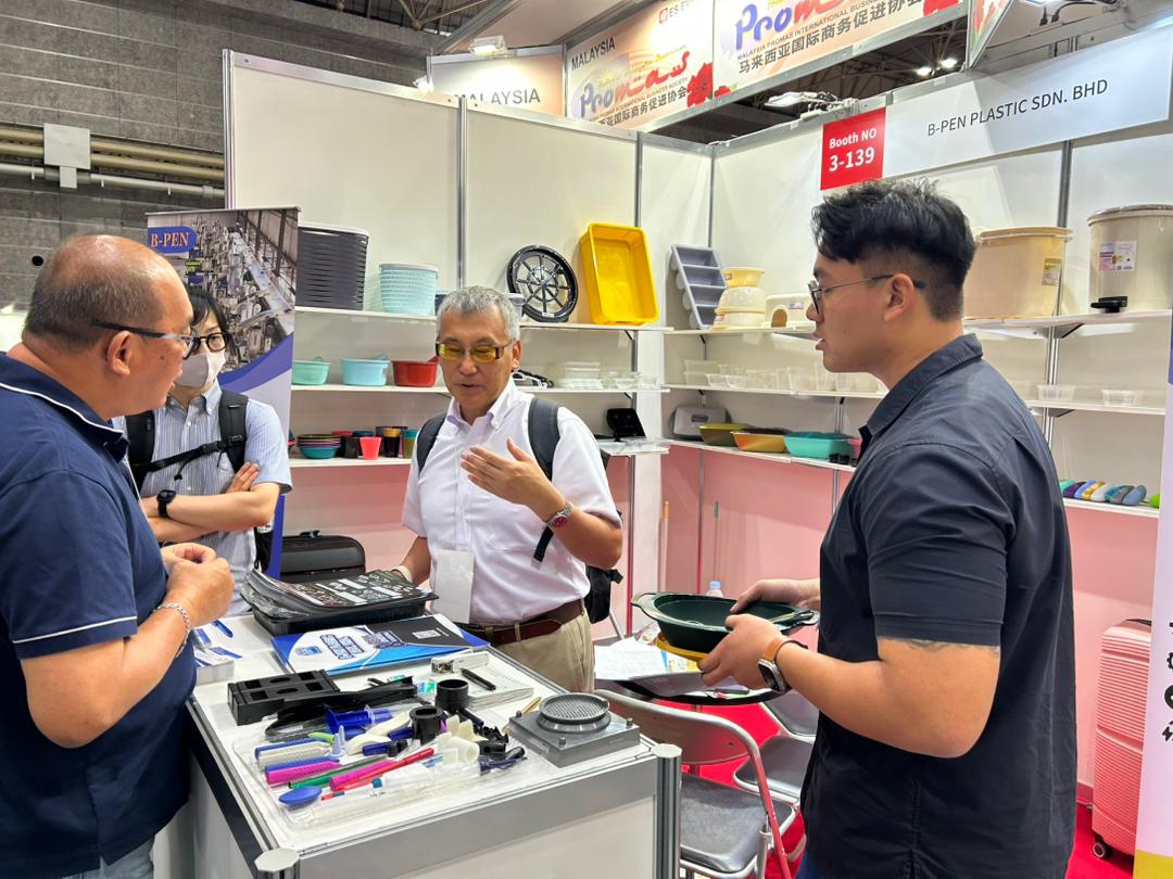 B-PEN team discussing molded samples with a Japanese visitor at the booth counter